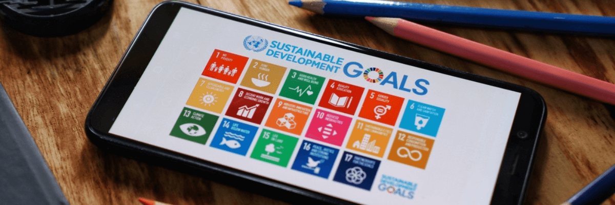 A imagem mostra um aparelho de celular exibindo na tela os Objetivos de Desenvolvimento Sustentável. No fundo, lápis de cor de cores variadas representam o impacto do trabalho deste tema na educação bilíngue.