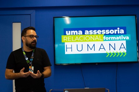A foto mostra um homem de camisa preta, com cabelo e barba bem aparados e usando óculos, enquanto gesticula durante uma apresentação de como o Be presta assessoria pedagógica para as escolas parceiras.