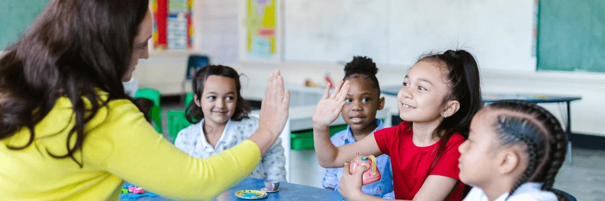 A imagem mostra uma professora bilíngue celebrando junto com um grupo de alunos um importante marco de aprendizado, ressaltando que o papel do professor bilíngue é muito além da mera avaliação de conteúdos.